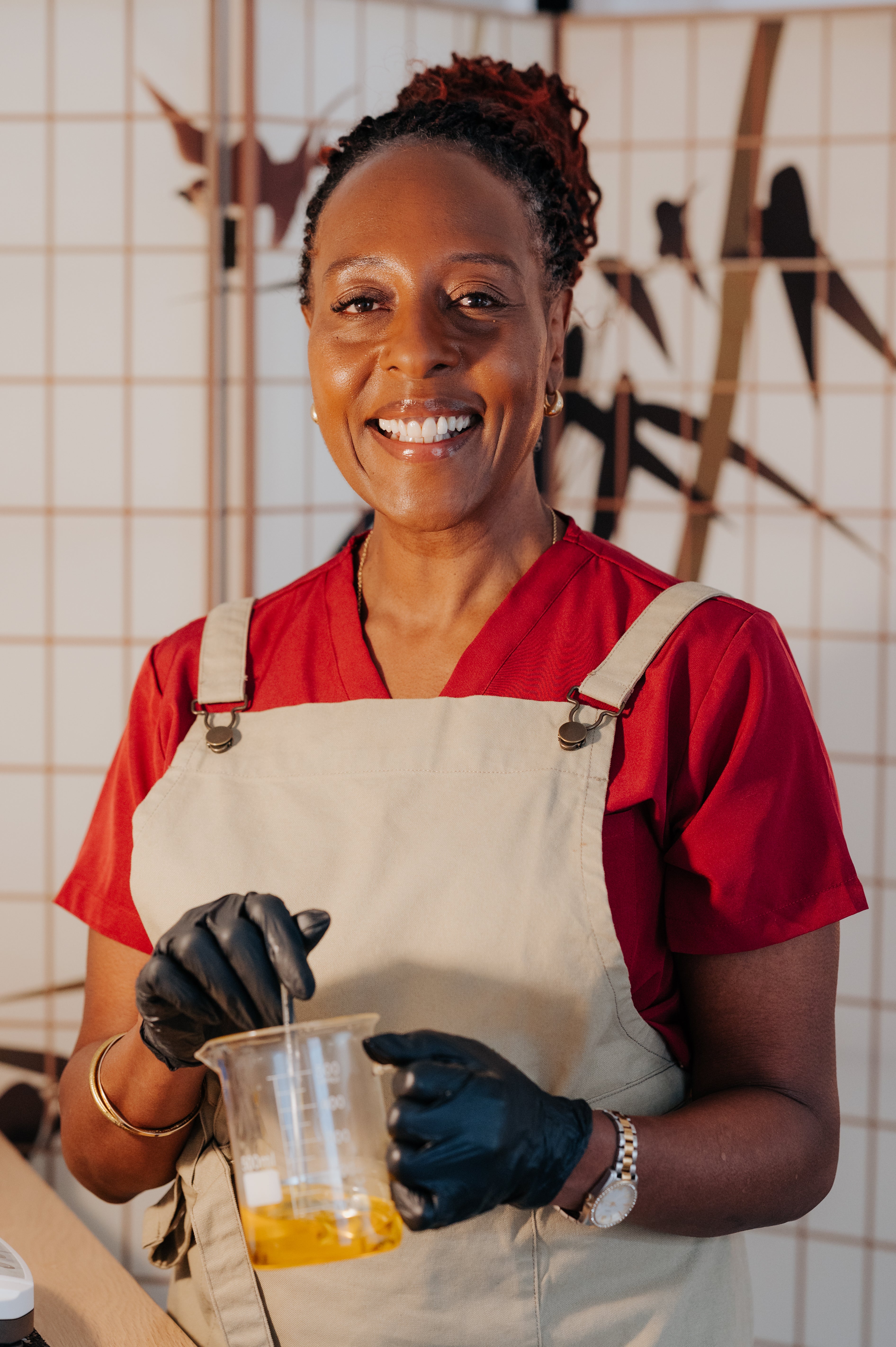 Equaa founder, Helen wearing a red shirt and beige apron holding a container mixing a golden oil substance, standing against a tiled wall with japanese bamboo pattern.